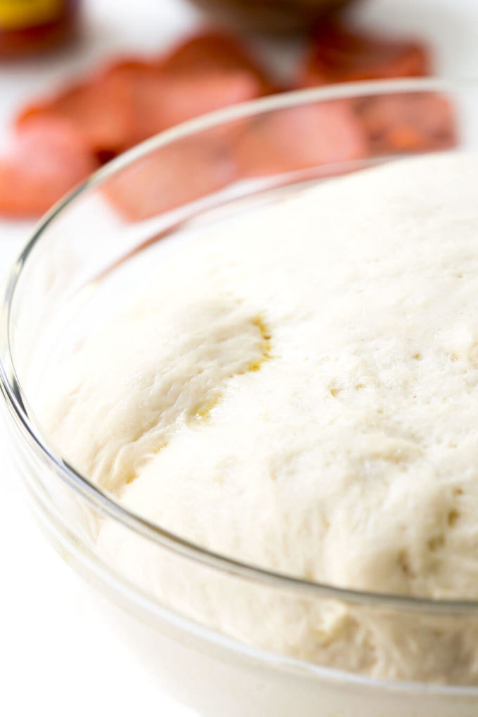 Rising homemade pizza dough in a glass mixing bowl.