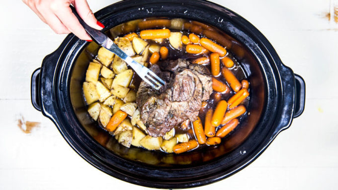 A chuck roast being held over a crock pot full of potatoes and carrots