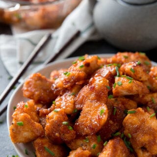 A bowl of baked orange chicken with chopsticks and a tea pot