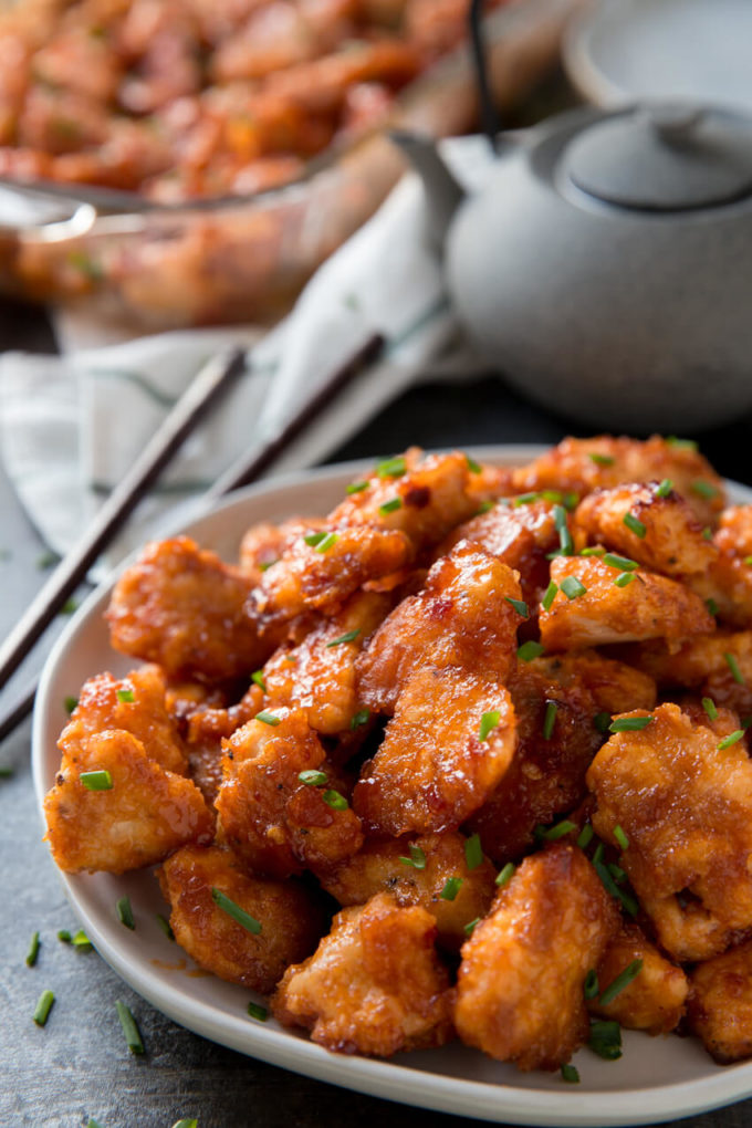 A bowl of baked orange chicken with chopsticks and a tea pot