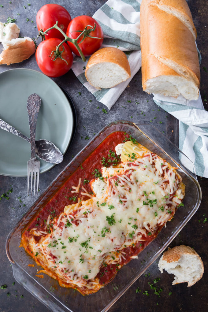 A glass baking dish of stuffed manicotti with French bread and fresh tomatoes