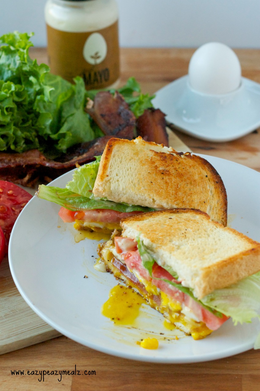 sandwich BELT on a white plate with lettuce, tomatoes, bacon, mayo and an egg in the background 