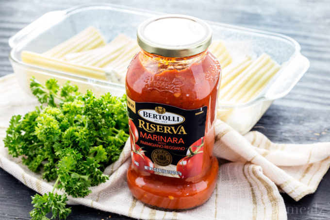 Bertolli pasta sauce in a jar on a dish towel with greens next to it and stuffed manicotti behind it