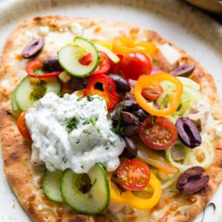 greek chicken flatbread on a cream colored plate. Topped with peppers, cucumbers, and other veggies.
