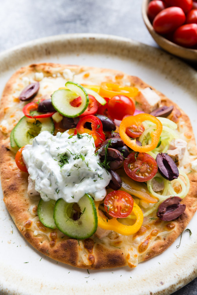 greek chicken flatbread on a cream colored plate. Topped with peppers, kalamata olives, cucumbers, tomatoes and cheese. With a sauce.