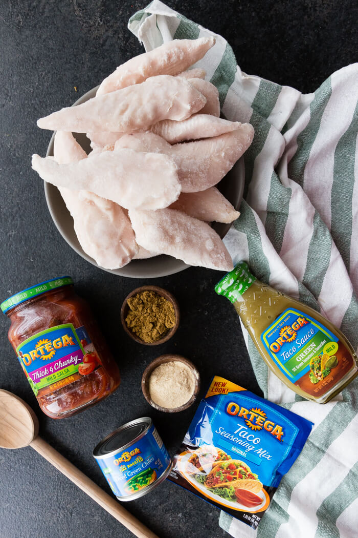 Frozen chicken in a bowl with mild salsa, mild taco sauce, taco seasoning, and green chilis around it with a wooden spoon on the table