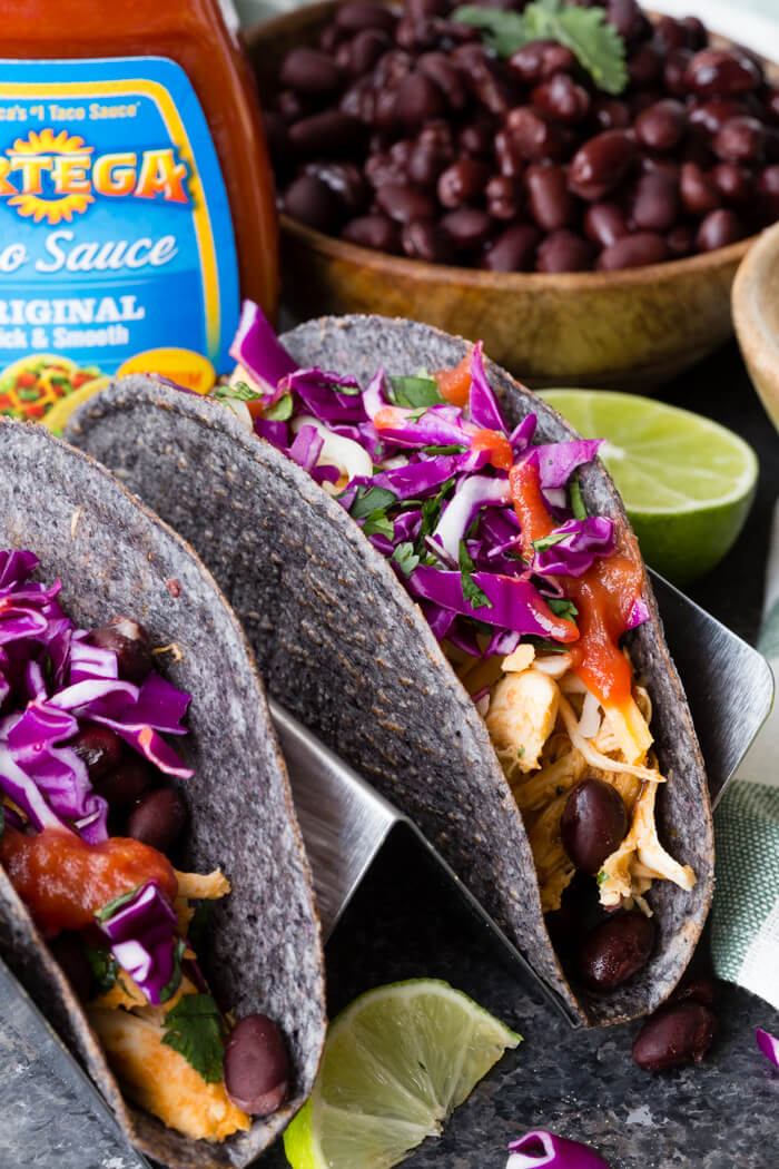 instant pot shredded chicken taco in a blue corn taco shell with black beans and taco sauce in the background