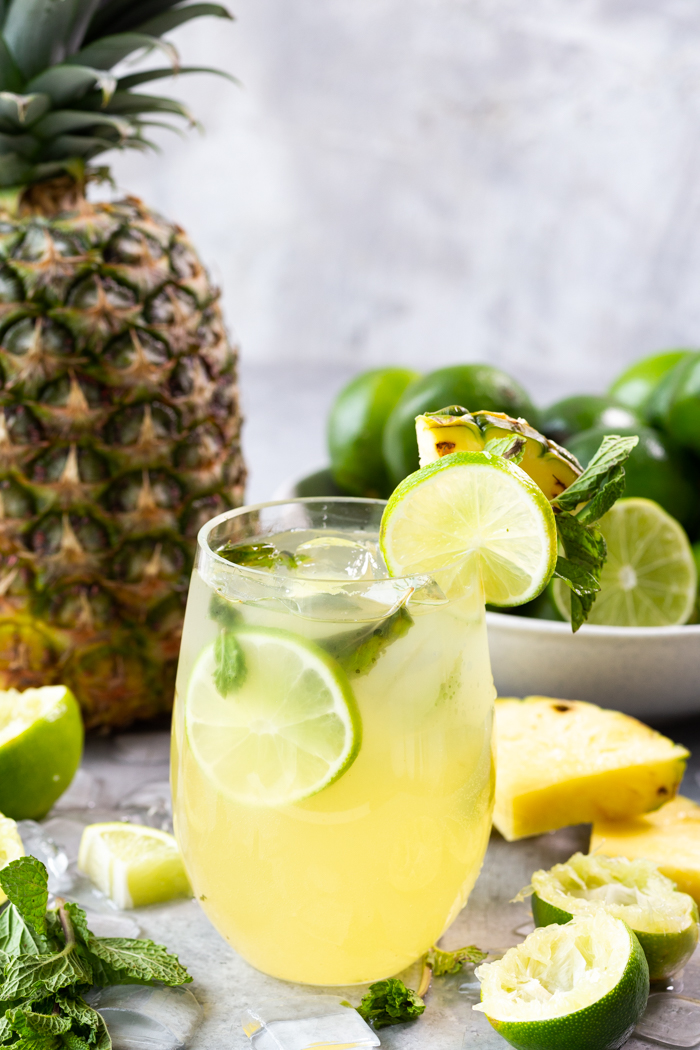 A glass of pineapple mojito with a pineapple and a bowl of limes in the background