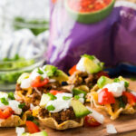 A cutting board with taco bites in Tostitos Scoops! a bag of chips in the background.