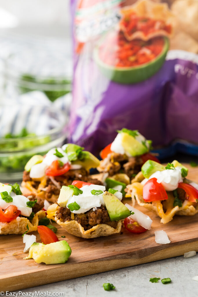 A cutting board with taco bites in Tostitos Scoops! a bag of chips in the background.