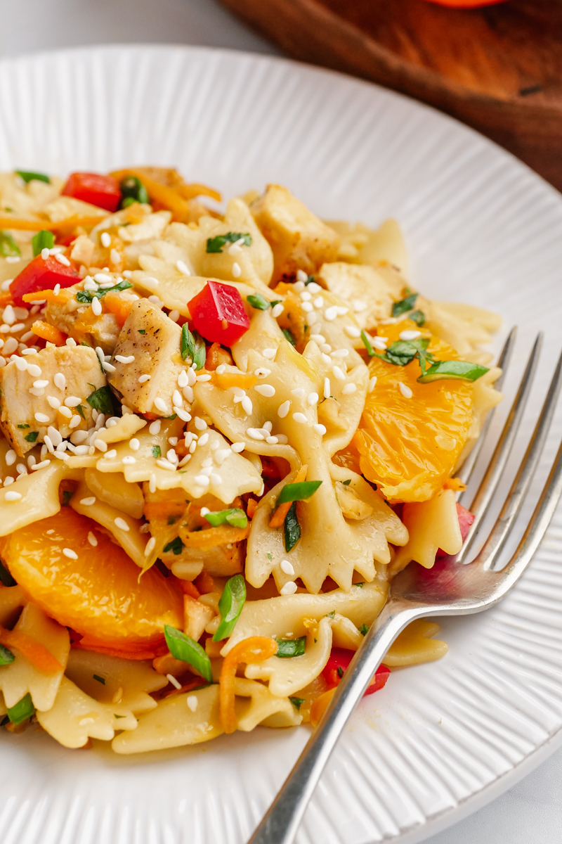 A white plate with a silver fork, pasta, mandarins, peppers, and green garnish. 