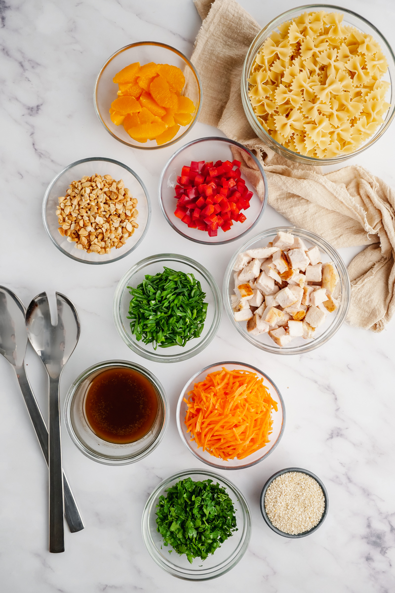 Small prep bowls with all the ingredients you need for Asian Pasta Salad, with utensils on the left side. 