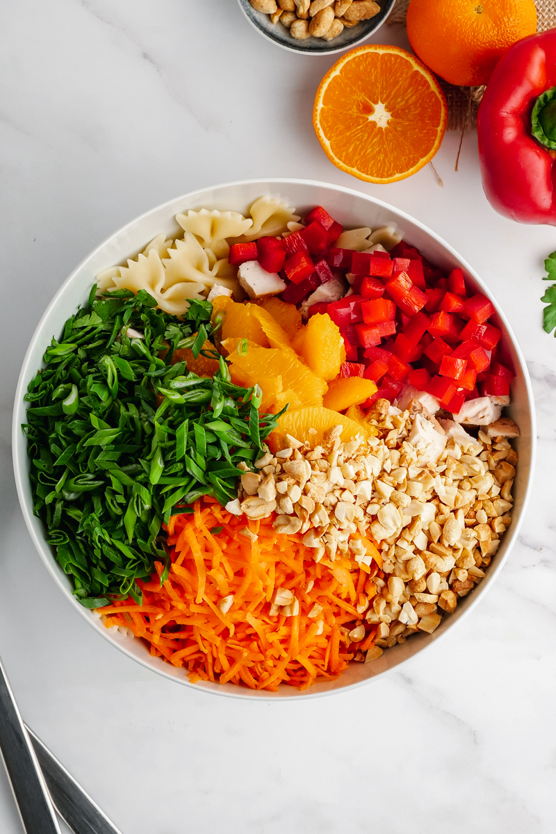 A white bowl with all the ingredients for Asian Pasta Salad, with oranges and a pepper on the top right hand corner. 