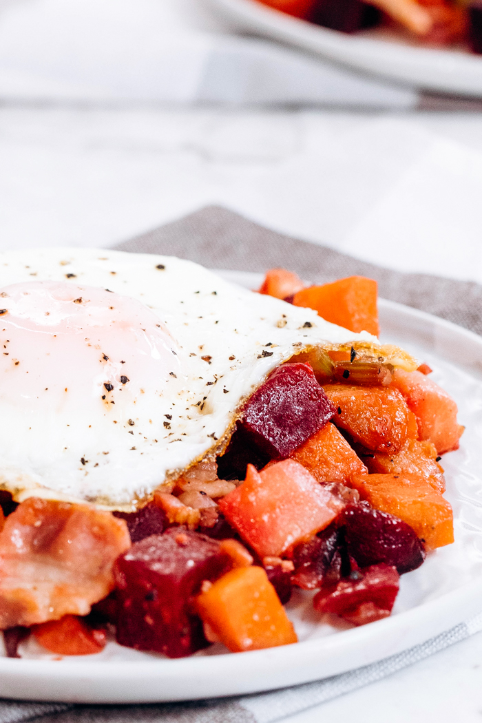 A white plate topped with veggie hash with bacon, and topped with a fried egg. 