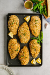 Breaded Chicken lying on cooking sheet, with lemons and herbs.