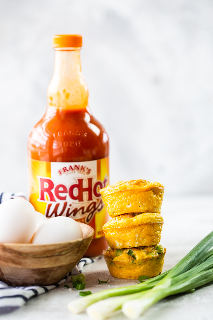 buffalo chicken egg muffins stacked on top of each other with whole eggs in a cup to the side. franks red hot wings sauce in the background