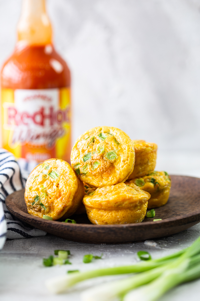 buffalo chicken egg muffins on a brown plate, with franks red hot blurry in the background. a bit of blue and white cloth showing.