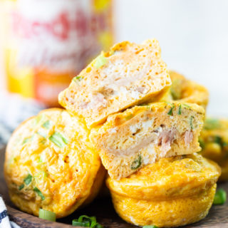 Brown plate with buffalo chicken egg muffins on top, one is cut in half, bottle of buffalo sauce in the background