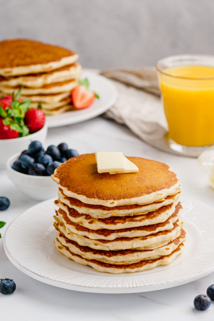 A stack of buttermilk pancakes homemade, on a white plate with orange juice in the background