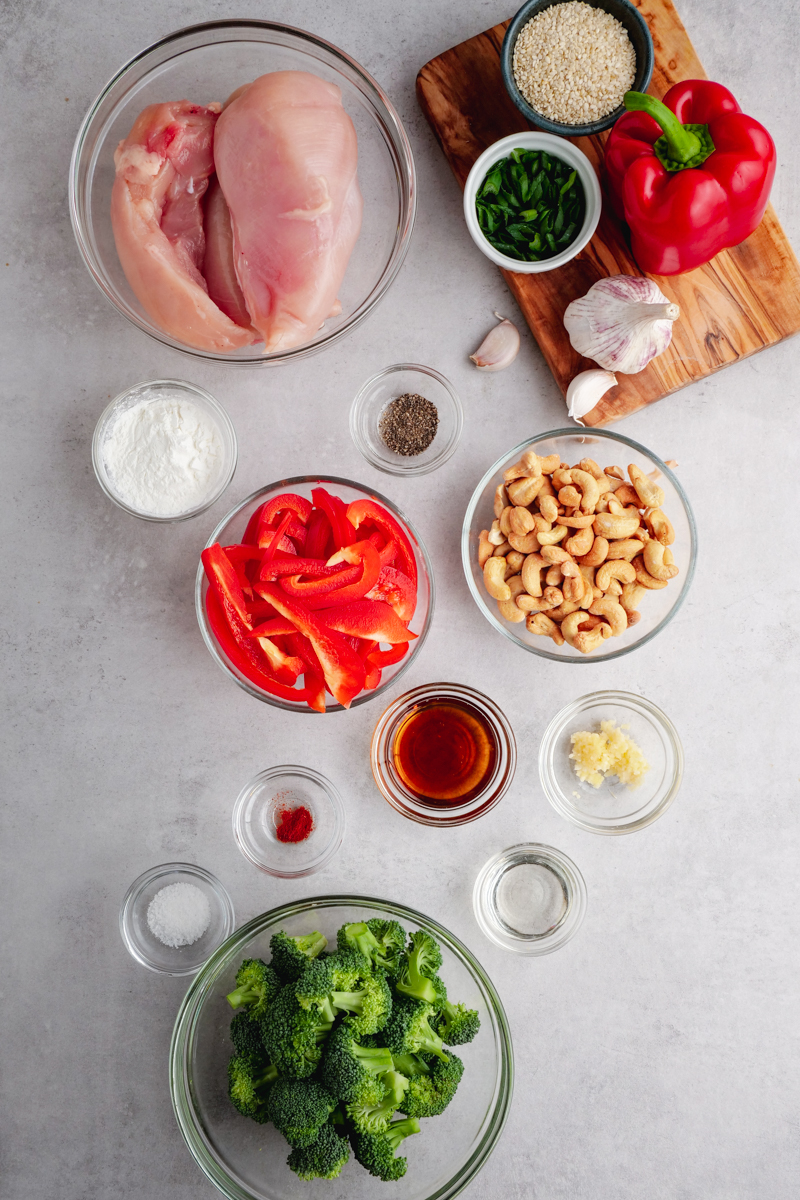 Portion cups with all the ingredients needed for cashew chicken, including raw chicken, vegetables, cashews. There is a cutting board in the top right hand cornder with more ingredients on top. 