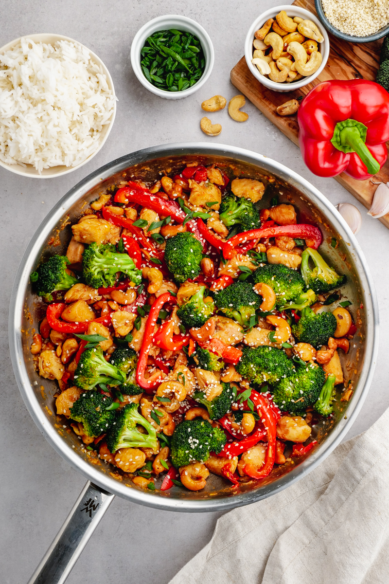 A stainless steel saute pan with cashew chicken inside, a bowl of rice, garnish, and a cutting board with cashews and red bell pepper on the sides. 