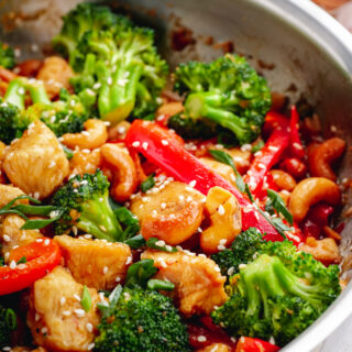 A stainless steel fry pan filled with broccoli, peppers, chicken, and cashew pieces, and topped with sesame seeds.