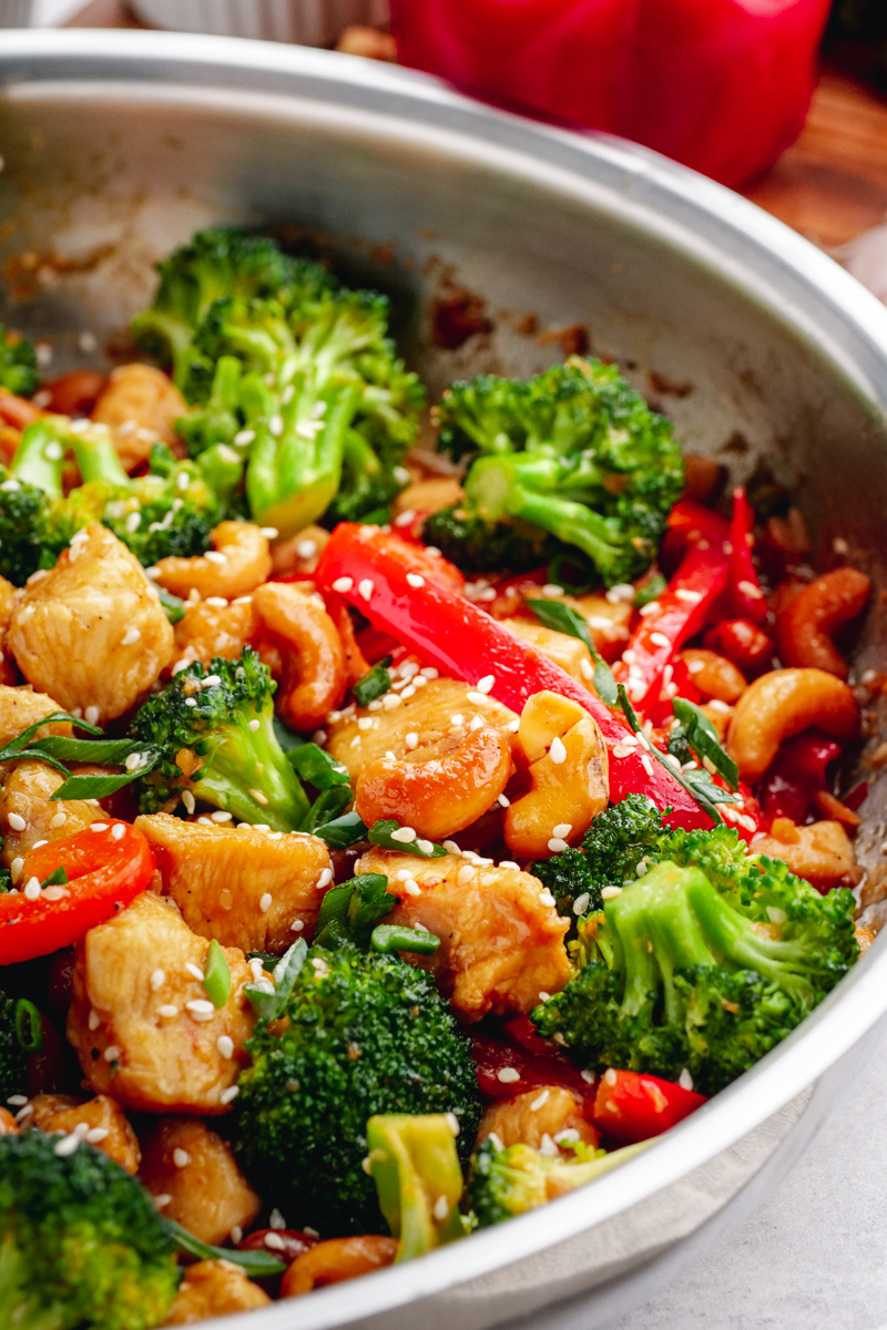 A stainless steel fry pan filled with broccoli, peppers, chicken, and cashew pieces, and topped with sesame seeds.