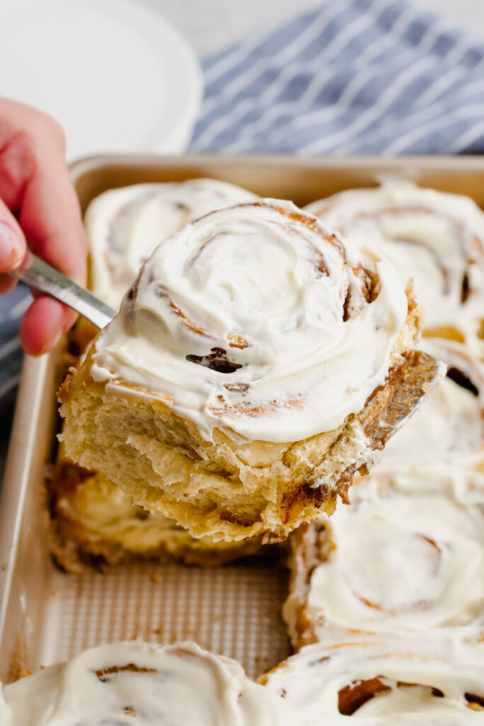 A pan of cinnamon rolls, taking a frosted cinnamon roll out of the pan.