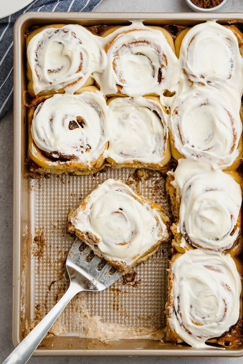 A pan of cinnamon rolls, some missing, a spatula with one frosted roll