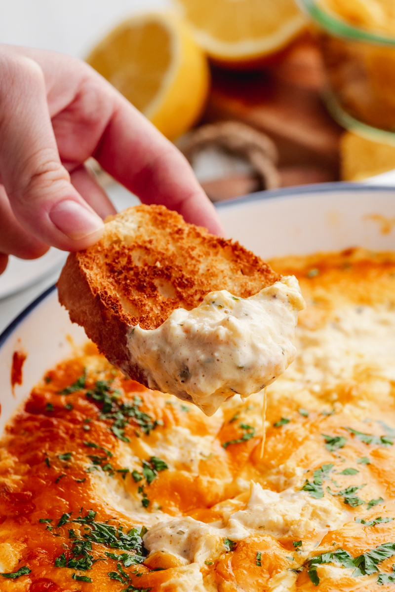 A white casserole dish loaded with creamy crab dip, and a hand with a slice of baguette dipped into the crab dip. 