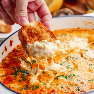 A white casserole dish of crab dip, with a baguette being dipped in, lemons in the background.