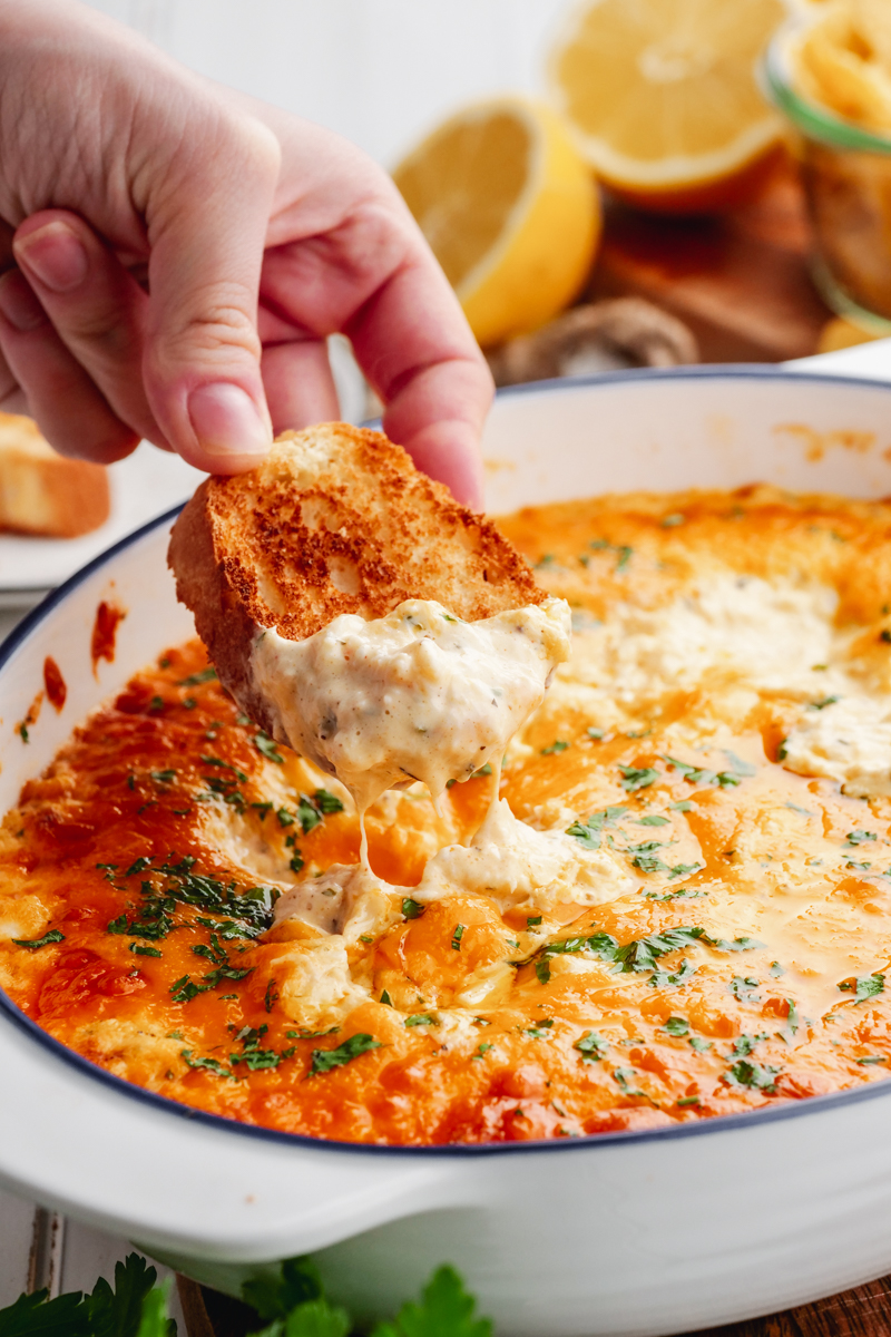 A white casserole dish of crab dip, with a baguette being dipped in, lemons in the background. 