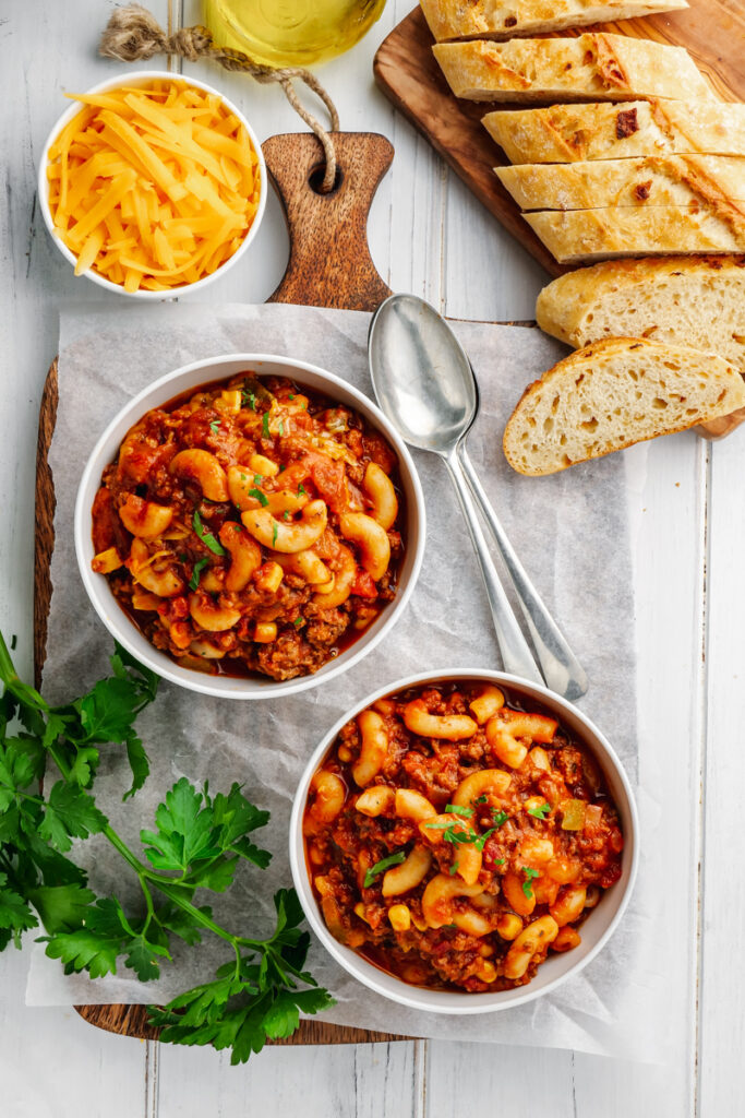 Two bowls of American Goulash, crusty bread on a cutting board, some green garnish and cheese. 