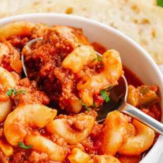 A bowl of American Goulash, white bowl, silver spoon, elbow macaroni, and some green garnish on top. Bread in the background.