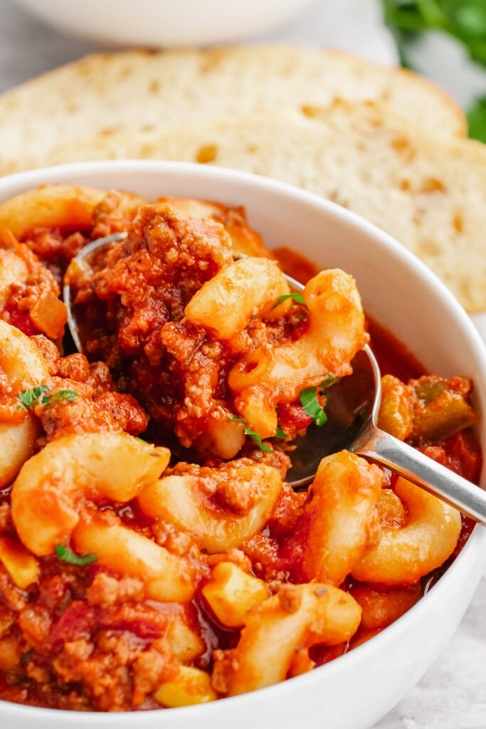 A bowl of American Goulash, white bowl, silver spoon, elbow macaroni, and some green garnish on top. Bread in the background. 