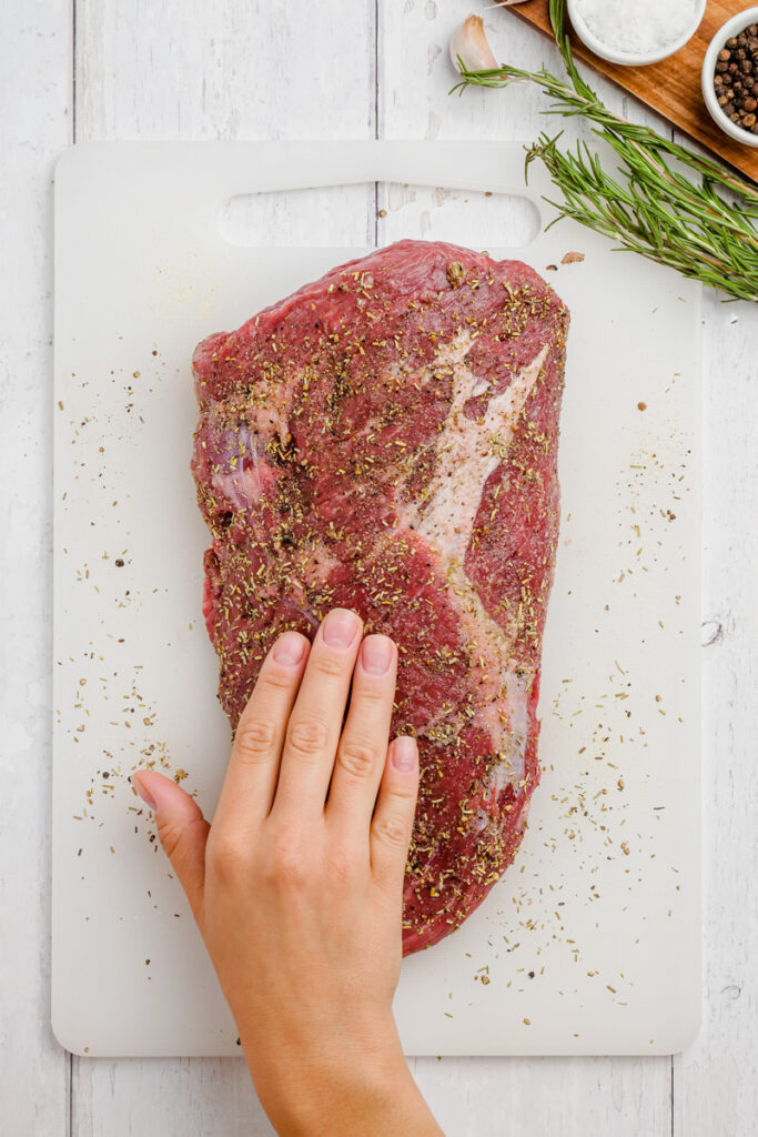 Instant Pot Sirloin Roast Beef on a white cutting board with spices all over it with basil to the side and a hand rubbing the spices around.