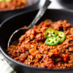 a black bowl full of keto chili, topped with jalapenos. This low carb chili has a spoon in it.
