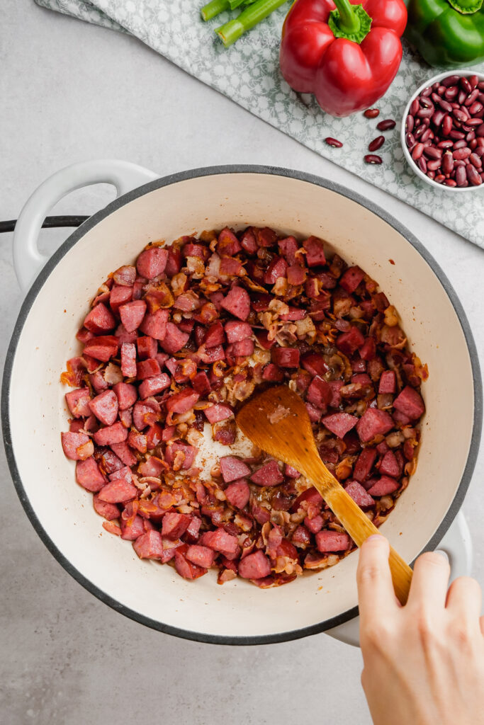 Red Beans and Bacon in a pot being stirred by a wooden spoon.