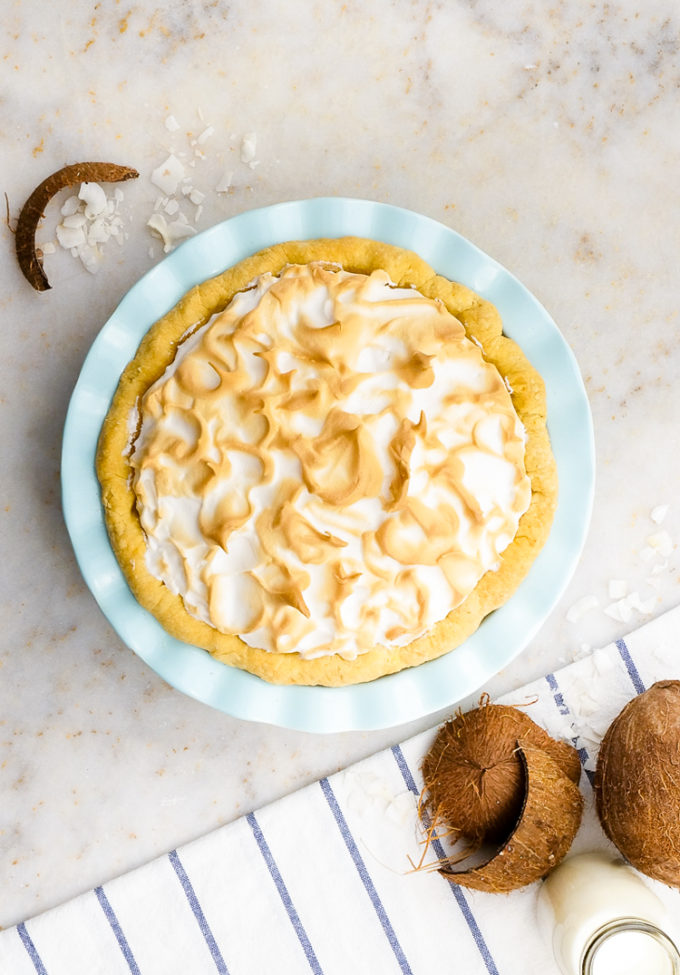 Deliciously meringue topped coconut cream pie with a thick rich coconut filling on a blue plate next to coconut shells