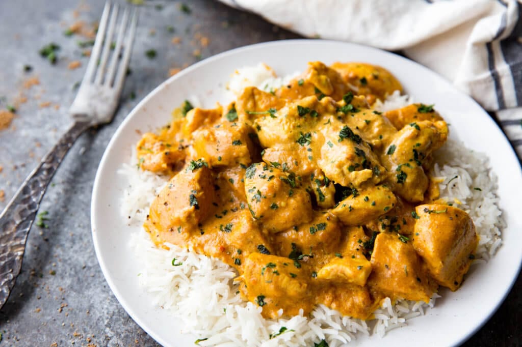 Indian butter chicken over white rice with cilantro as a garnish on a white plate with a silver fork 