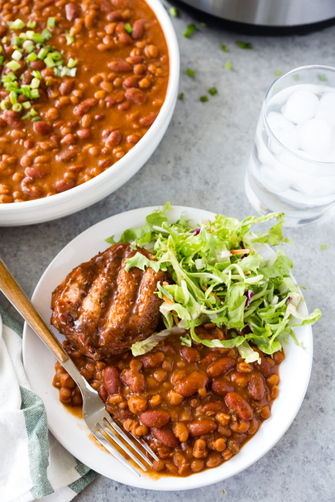Summer staple, instant pot baked beans with a pork chop and green salad, on a white plate with a white bowl of beans 