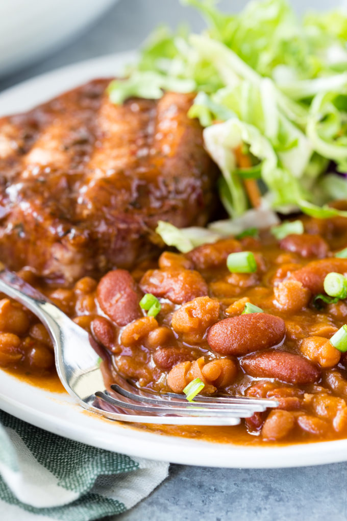 Delicious and easy to make Baked beans, a white plate with baked beans, a pork chop, and greek salad, fork on the left side. 