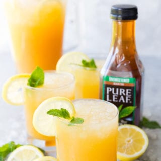 A few glasses of Mango Black Tea Lemonade with a lemon and mint garnish, and a pitcher of mango black tea lemonade in the background, as well as a bottle of Pure Leaf black tea on the side.