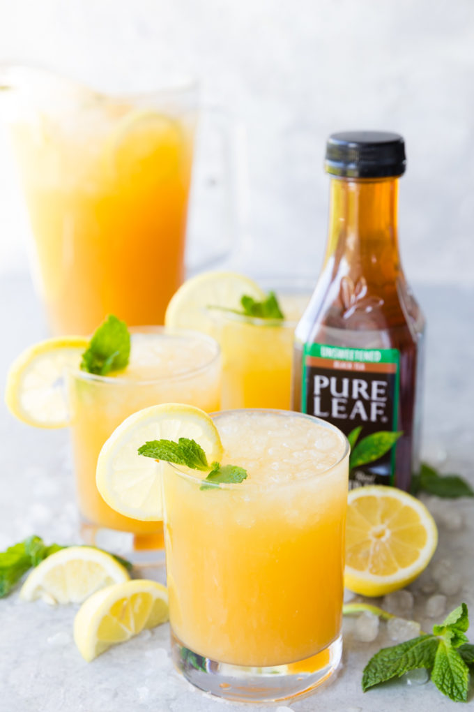 A few glasses of Mango Black Tea Lemonade with a lemon and mint garnish, and a pitcher of mango black tea lemonade in the background, as well as a bottle of Pure Leaf black tea on the side.