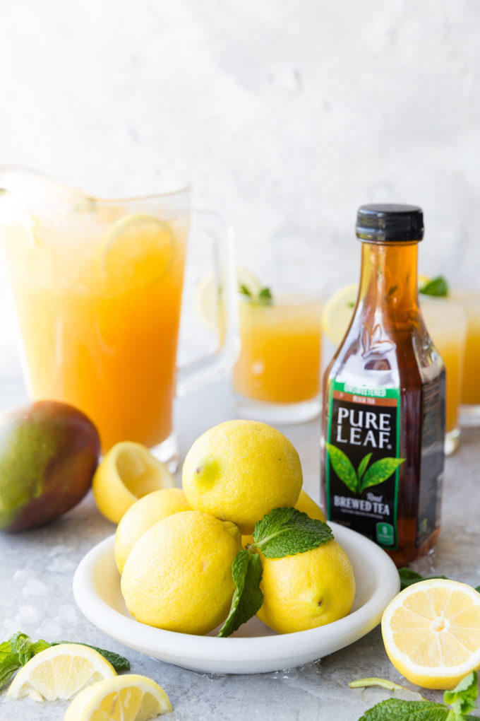 A white plate with lemons and mint, a bottle of pure leaf black tea, a mango, and a pitcher of Mango Black Tea Lemonade