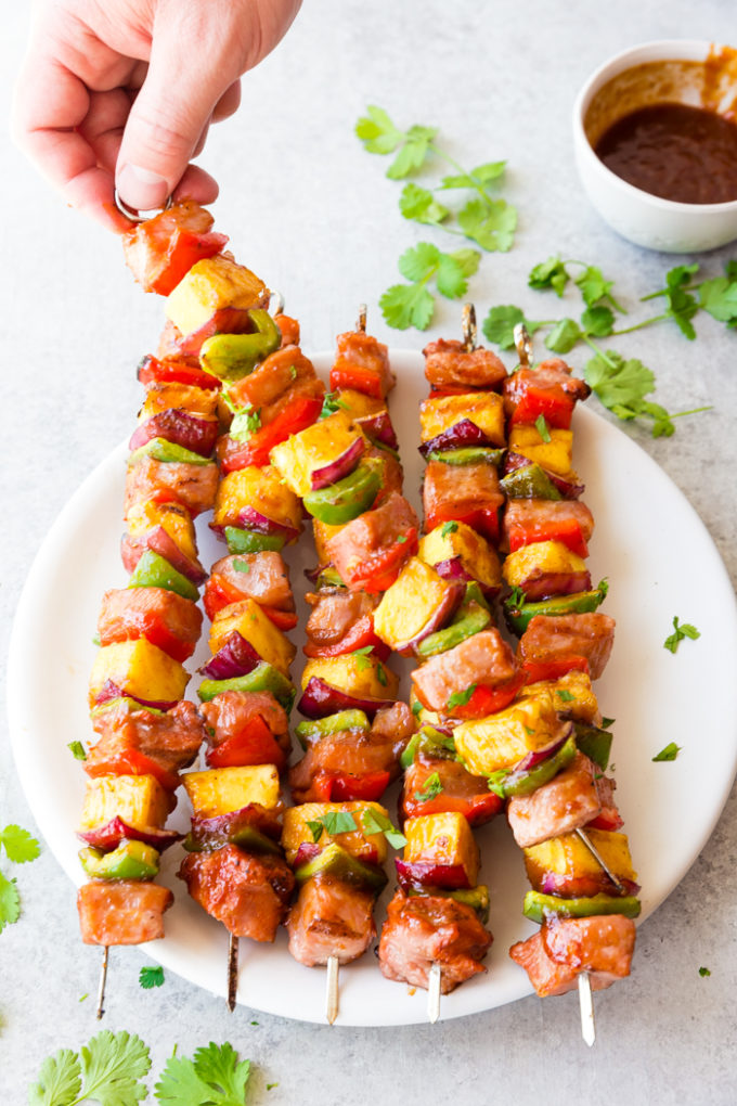 A white plate with 6 sticky bbq pork skewers cilantro, and bbq sauce for dipping