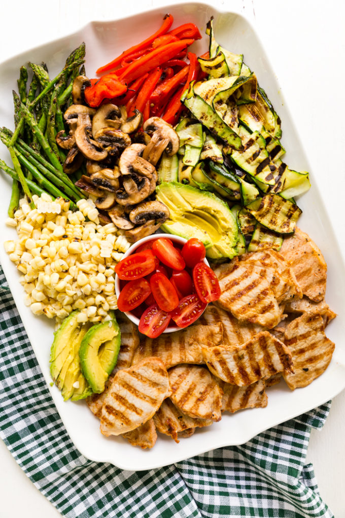 A platter of grilled pork, avocado, corn, tomatoes, asparagus, mushrooms, red pepper, and zucchini for grilled pork power bowls