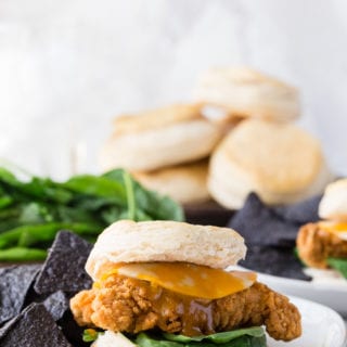 A buttermilk biscuit sandwich with cheese, spinach, and honey mustard sauce on a white place with blue corn tortilla chips