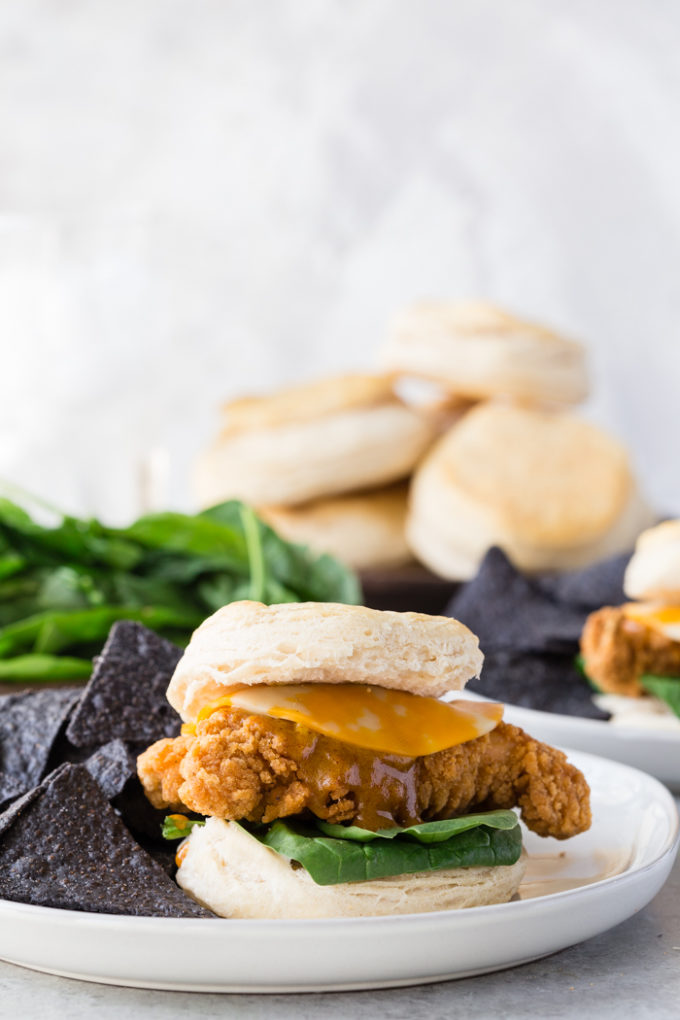 A buttermilk biscuit sandwich with cheese, spinach, and honey mustard sauce on a white place with blue corn tortilla chips