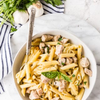 Roasted garlic and herb penne pasta in a white bowl with a blue and white napkin, cloves of garlic, and fresh parsley